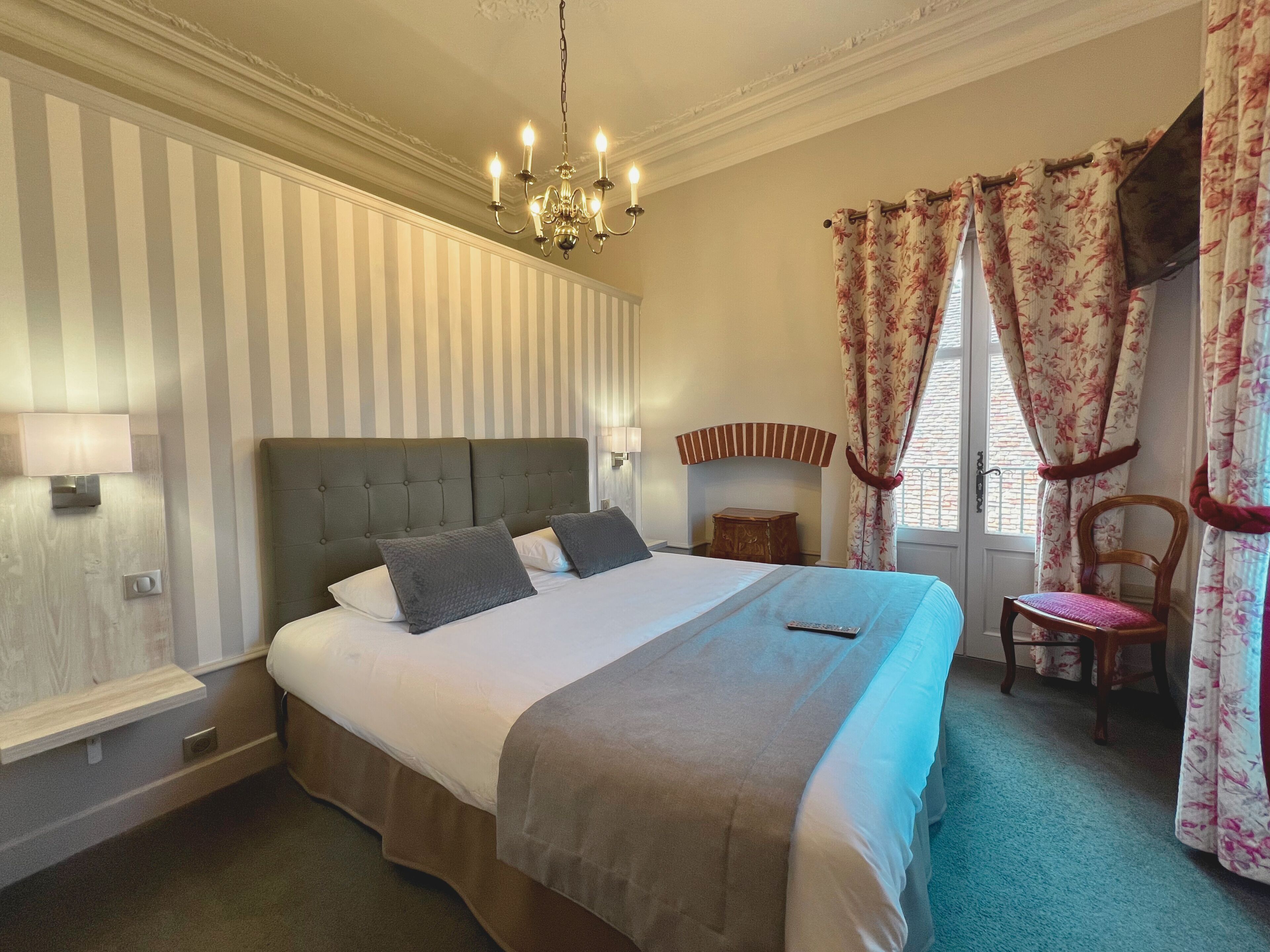 A neatly arranged bedroom with a large bed, a bedside table, a chair, a chandelier, and curtains.