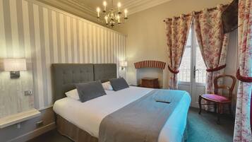 A neatly arranged bedroom with a large bed, a bedside table, a chair, a chandelier, and curtains.