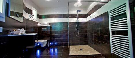 A modern bathroom with a glass-enclosed shower, dark tiled walls, a white towel rack, and a visible toilet.