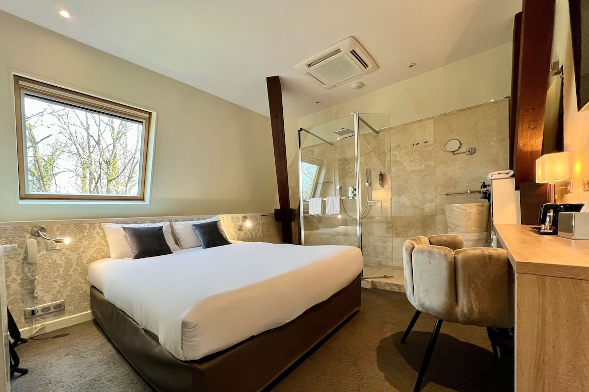 A modern hotel room with a large bed, a glass-enclosed shower, a desk with a lamp, and a view of trees through a window.