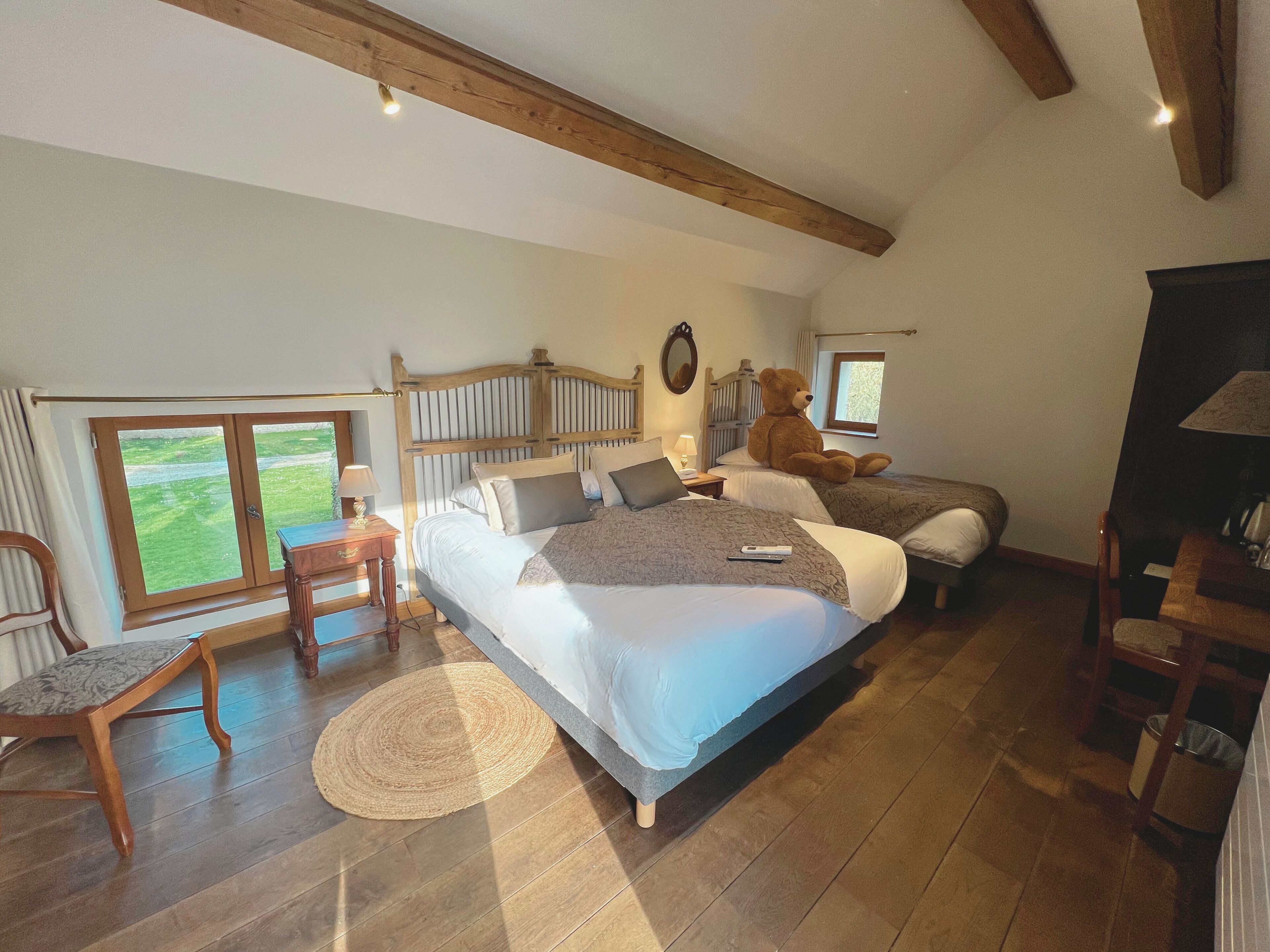 Une chambre avec un grand lit, un plus petit lit, une chaise, un bureau et une fenêtre donnant sur un paysage verdoyant.
