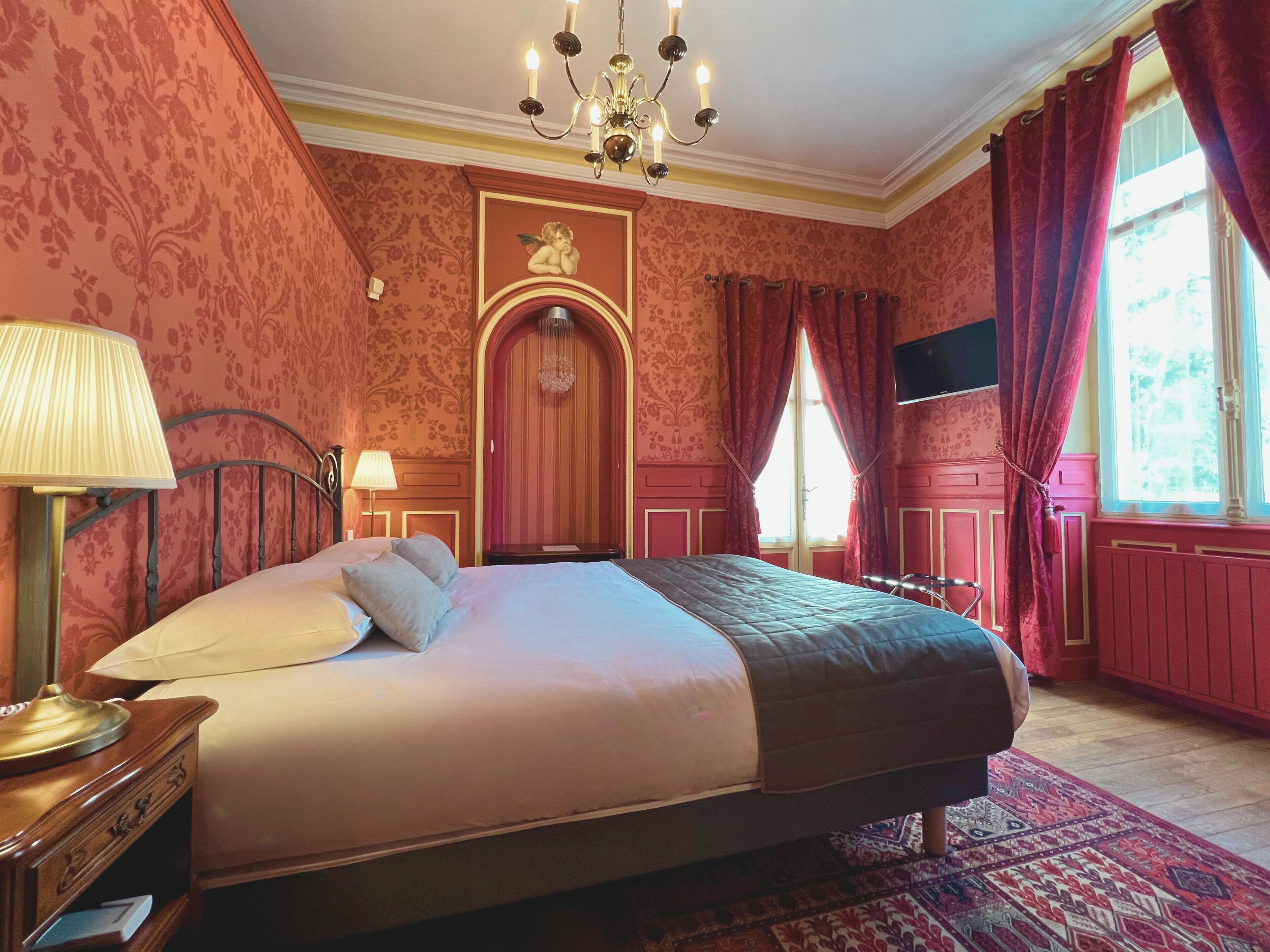 Une chambre à coucher avec un grand lit, un lustre, une table de chevet en bois et un mur décoré d’un motif rouge.