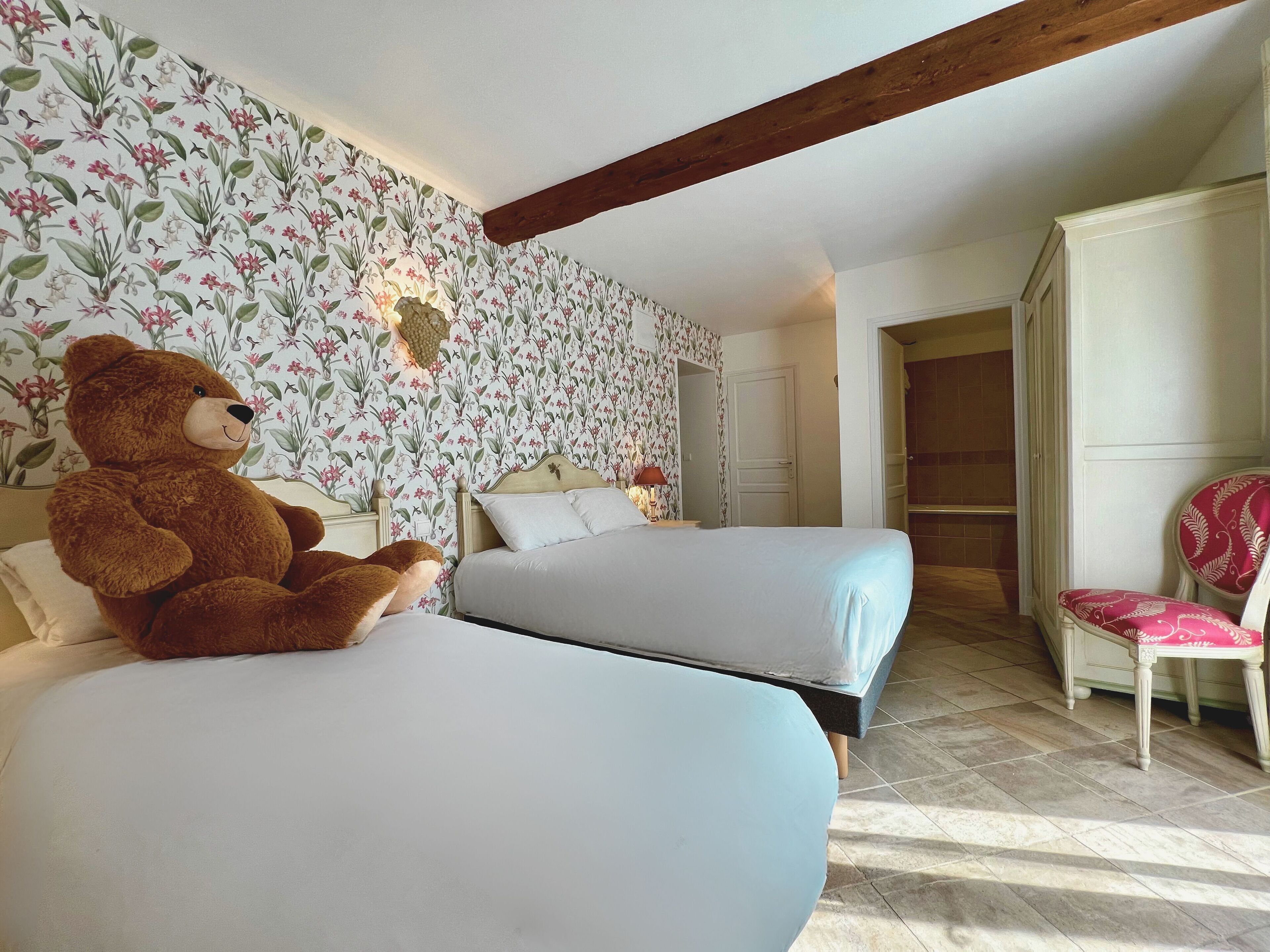 Een hotelkamer met twee bedden, een grote knuffelbeer op een van de bedden, bloemenbehang, een houten balk in het plafond en een stoel met een rood-wit patroon.