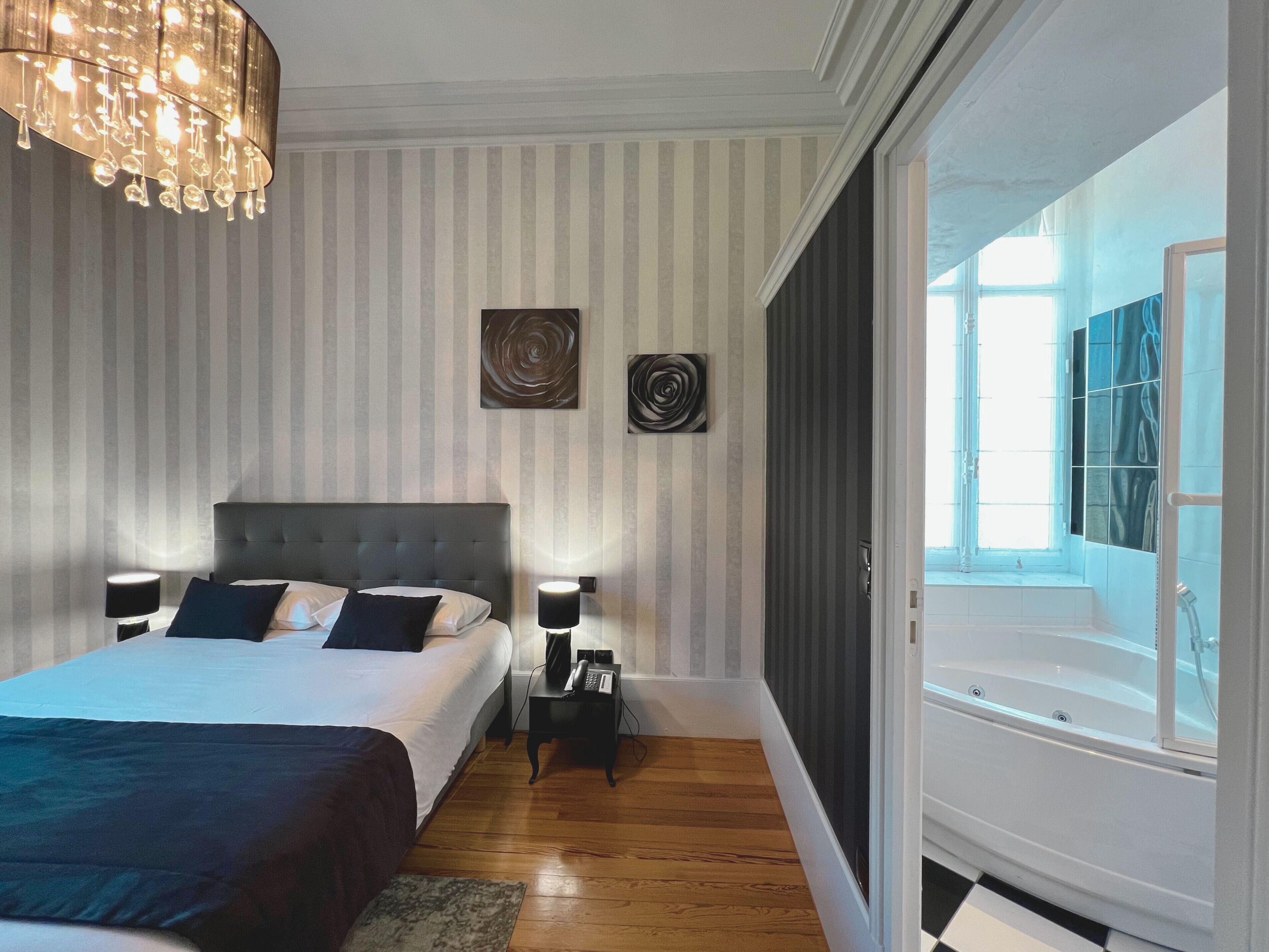 A modern bedroom with a large bed, bedside lamps, a chandelier, and a view of a bathroom with a bathtub.