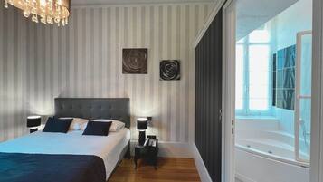 A modern bedroom with a large bed, bedside lamps, a chandelier, and a view of a bathroom with a bathtub.