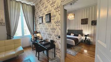 A room with a desk, chair, and a bed with a headboard. There is a window with curtains, a radiator, and a chandelier.