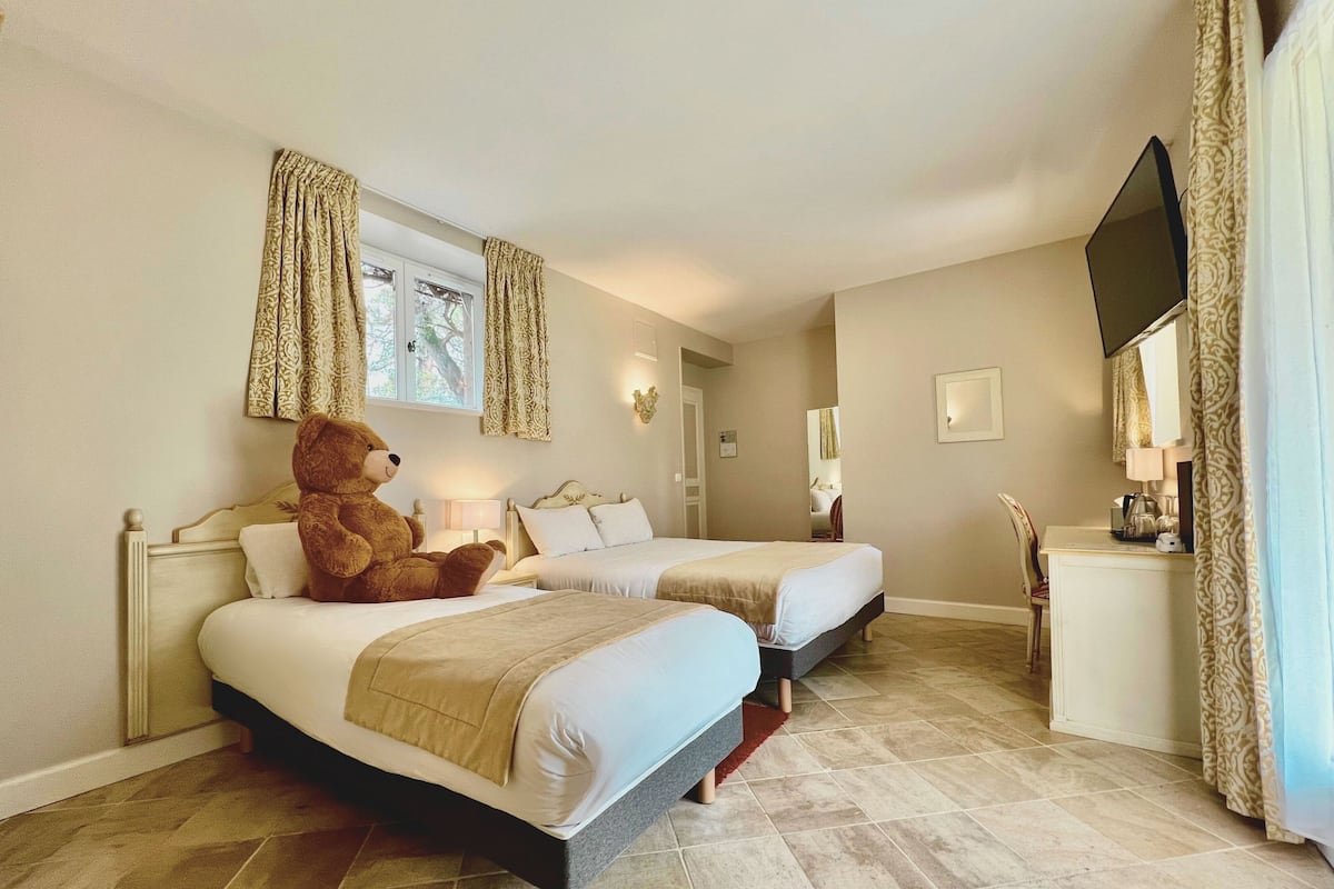 A hotel room with two beds, a large teddy bear on the left bed, a desk with a lamp, and a mirror on the right wall.