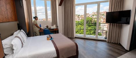 A hotel room with a bed, a desk, a chair, a TV, and a large window with a city view.