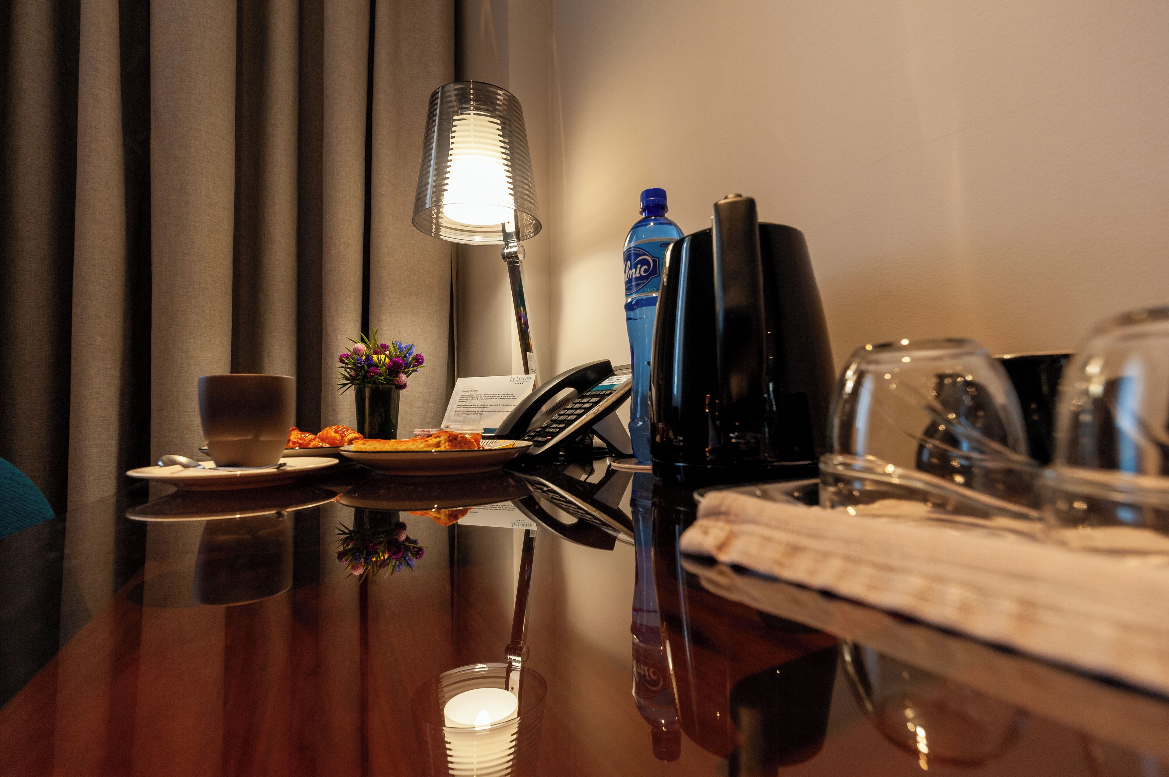 Une chambre d’hôtel avec un bureau en bois, une lampe, un téléphone, une bouteille d’eau et un verre.