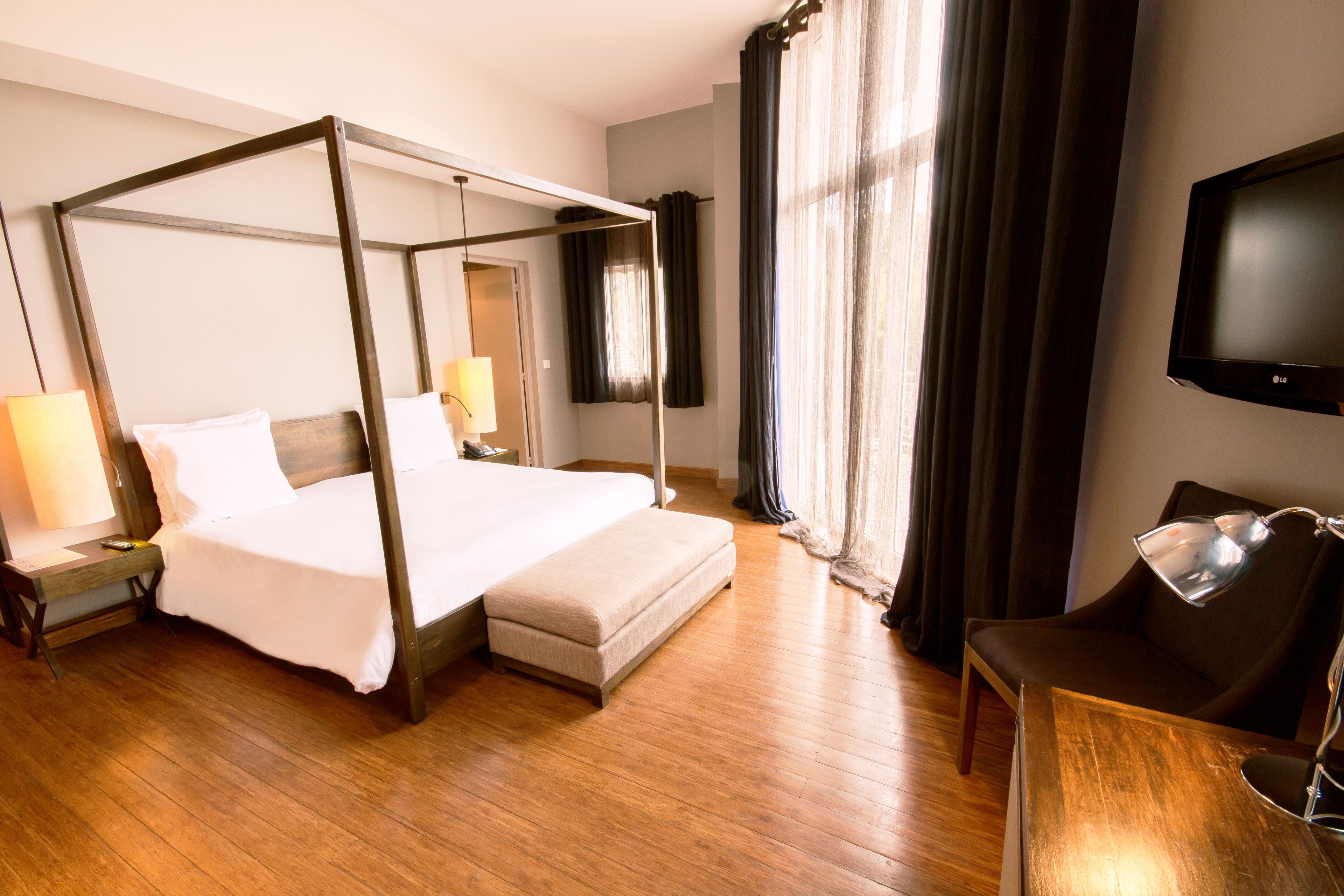 A four-poster bed with a wooden frame, a bedside table with a lamp, a TV mounted on the wall, and a chair with a desk.