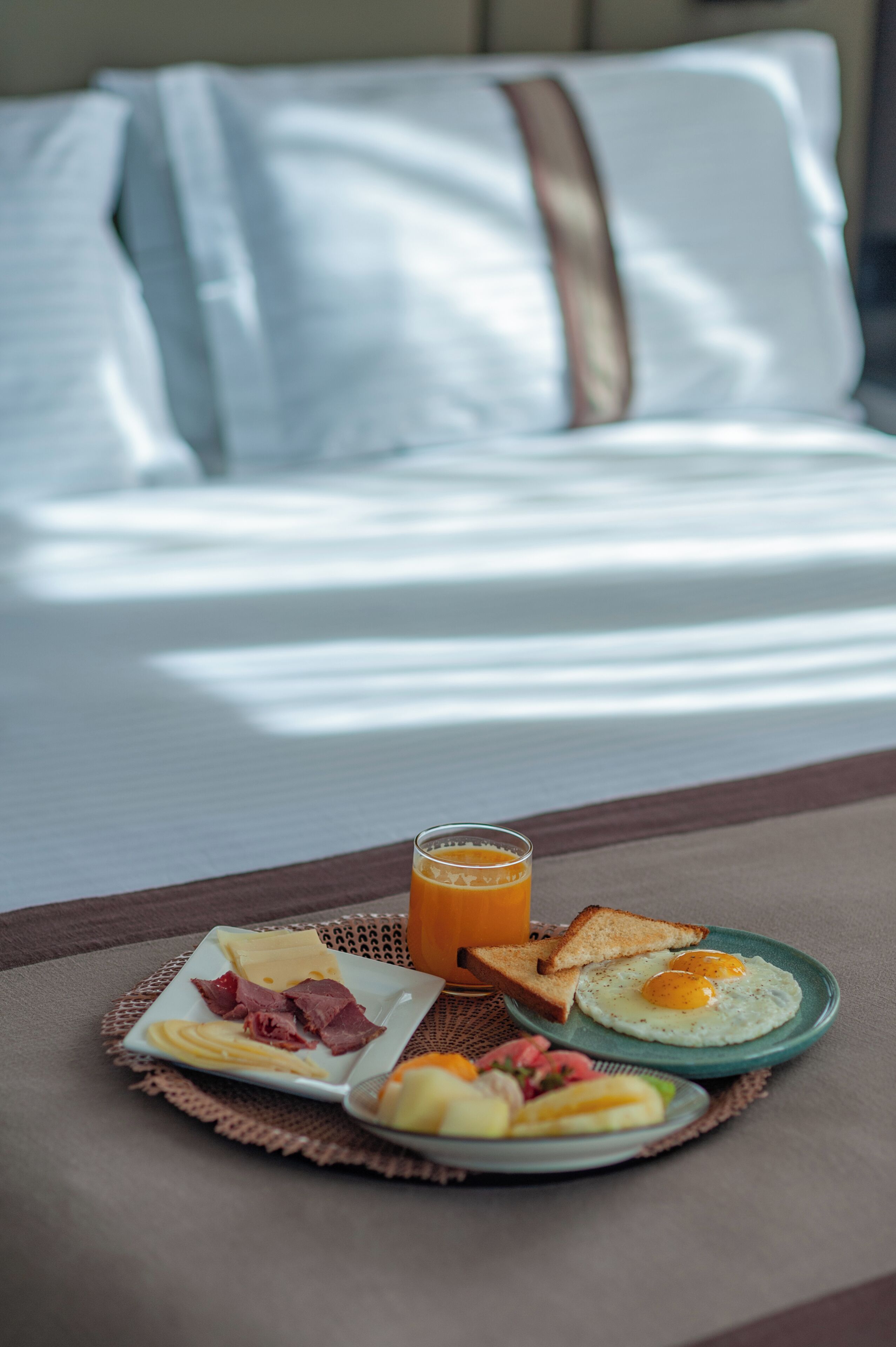 A breakfast setting with a plate of eggs, toast, and ham, a glass of orange juice, and a bed in the background.