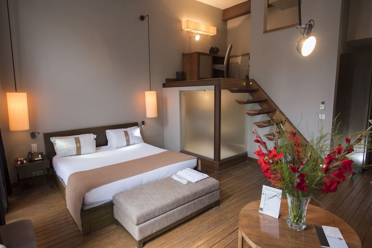 A hotel room with a large bed, a wooden bench, a desk with a vase of flowers, a wall-mounted light, and a staircase.