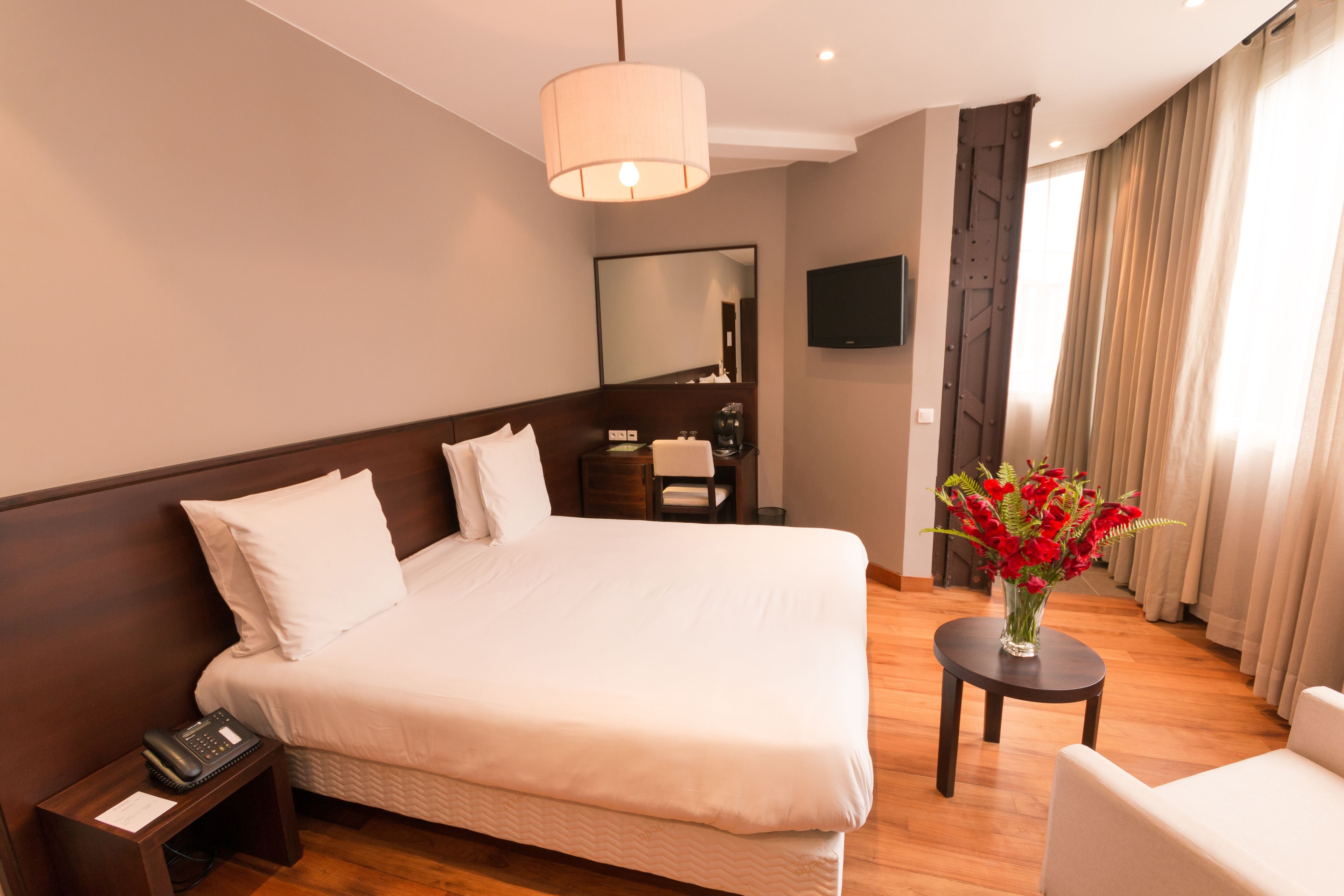 A hotel room with a large bed, a bedside table with a phone, a small table with a vase of flowers, a flat-screen TV mounted on the wall, and a large window with curtains.