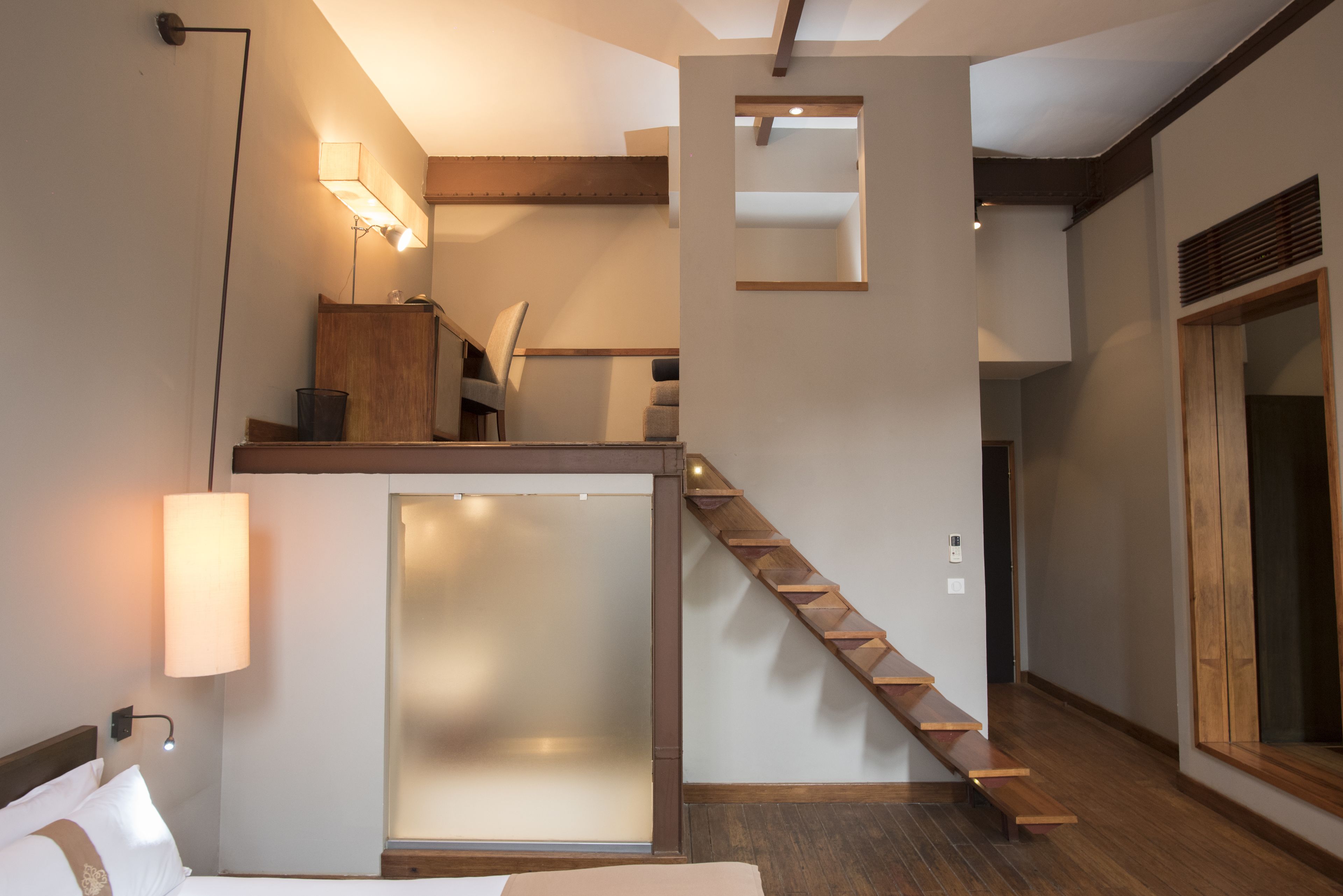 A modern hotel room with a wooden staircase, a large window, and a bed with a nightstand.