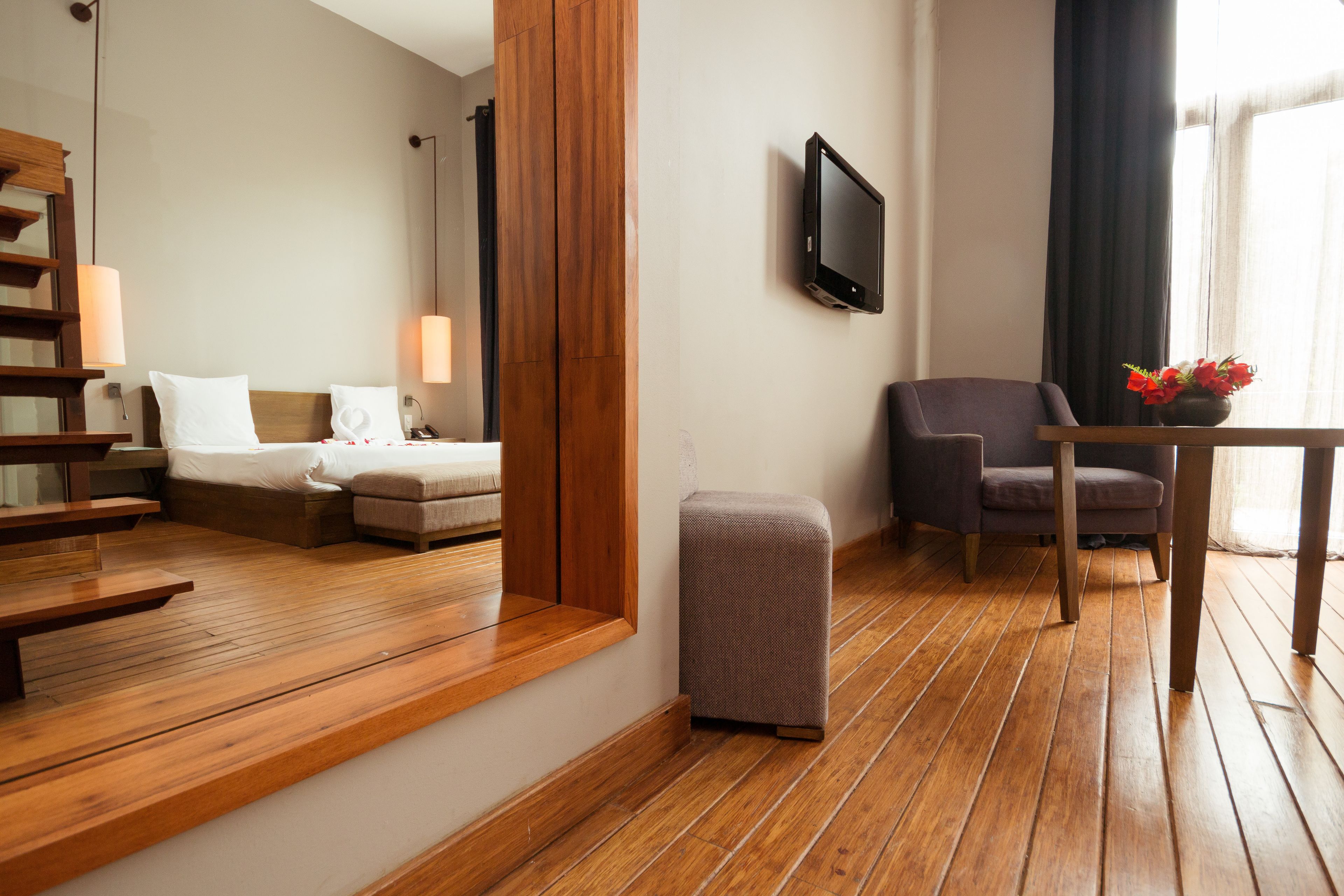 A hotel room with a wooden floor, a bed, a chair, a small table with a vase of flowers, and a television mounted on the wall.