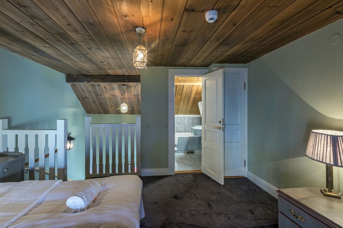 Un dormitorio con techo de madera, una cama con ropa blanca, una mesita de noche, un aparador con una lámpara y vistas al baño.