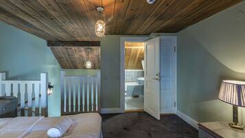 A bedroom with a wooden ceiling, a bed with white linens, a nightstand, a dresser with a lamp, and a view into the bathroom.