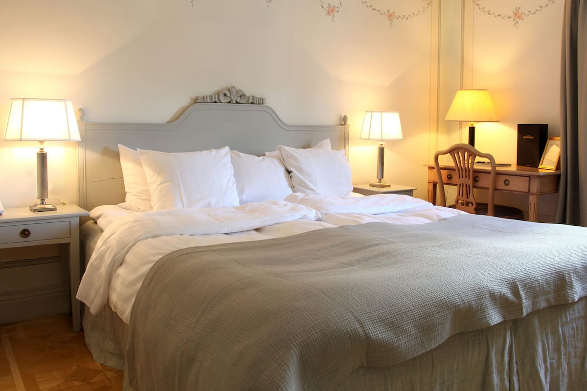 Una cama bien tendida con sábanas blancas, dos mesitas de noche con lámparas, una silla de madera y un escritorio con una lámpara.