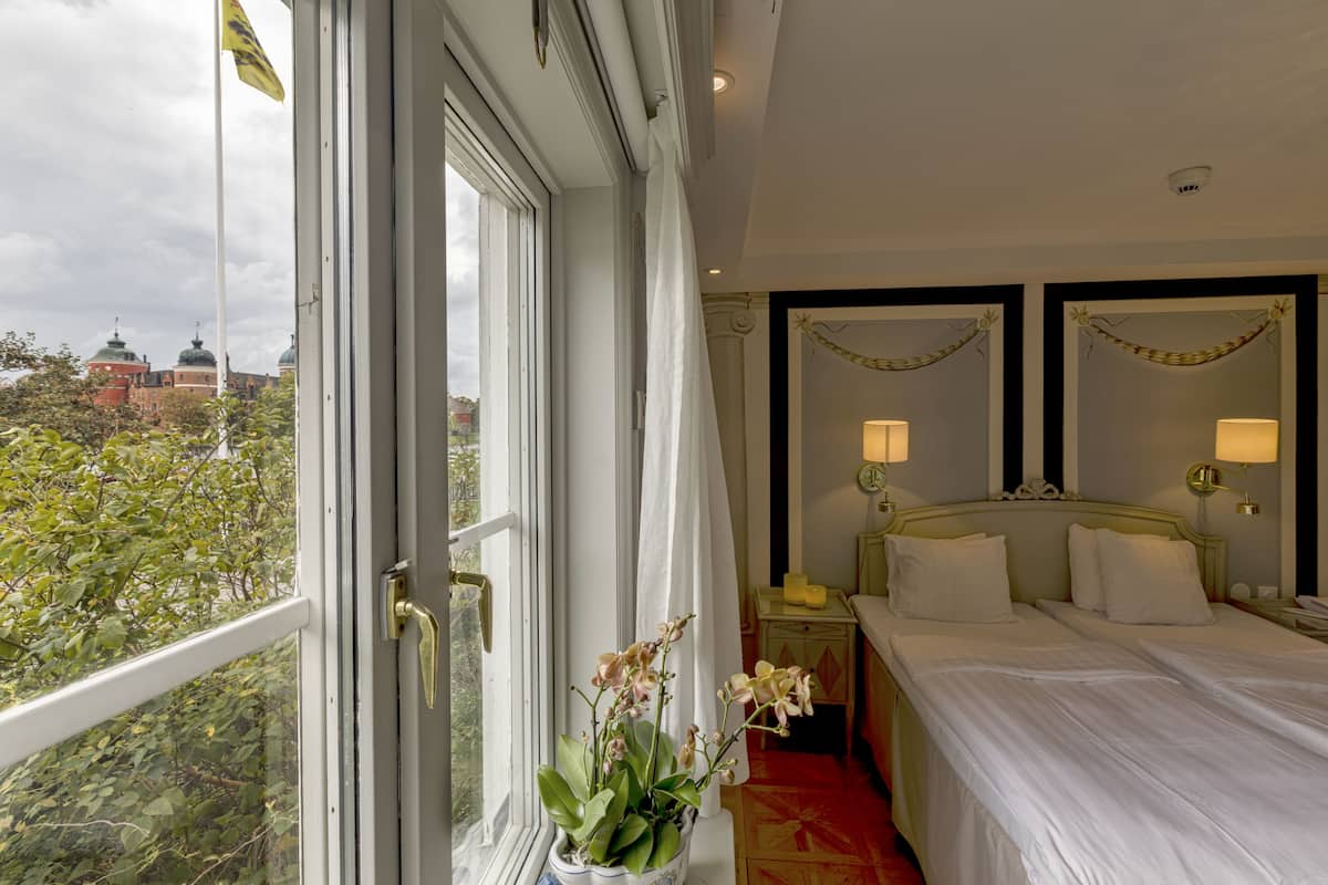 Habitación de hotel con un ventanal grande, una cama con ropa blanca, dos lámparas de noche y una mesita con lámpara.