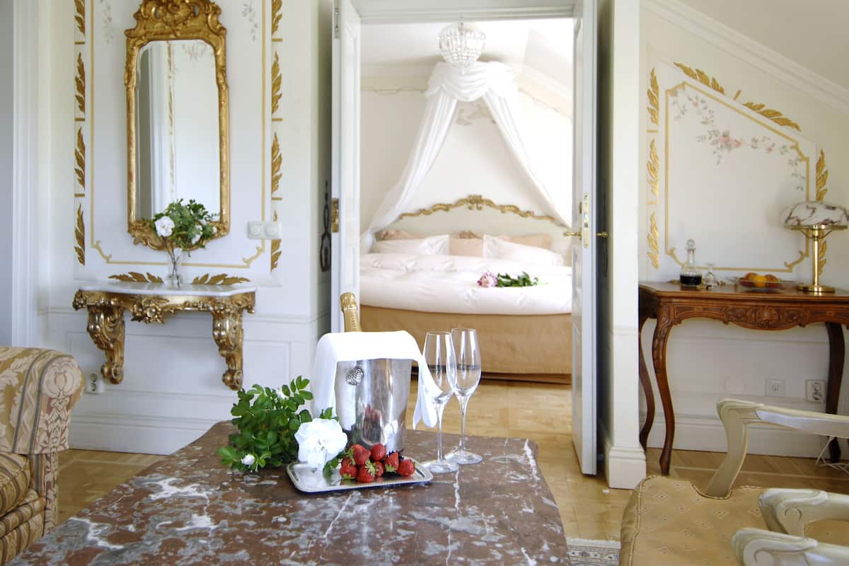 Un dormitorio lujoso con cama con dosel, una mesita de mármol con una botella de champán, copas y fresas, y un espejo con marco dorado.