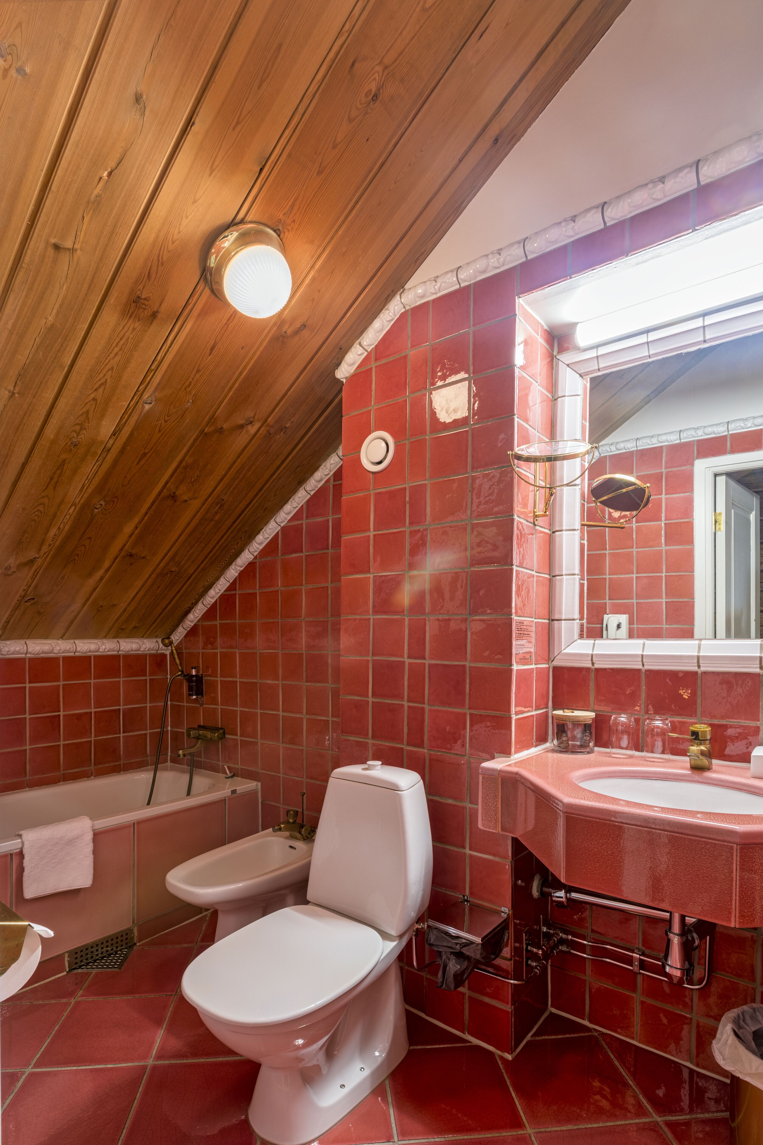 Een badkamer met een houten plafond, rode betegelde muren, een wit toilet, een bidet, een badkuip en een wastafel met spiegel.