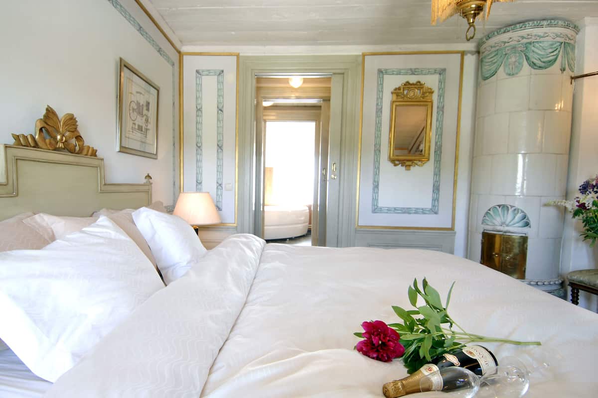 Habitación de hotel con una cama grande, una mesita de noche con lámpara, un espejo y un florero con flores.