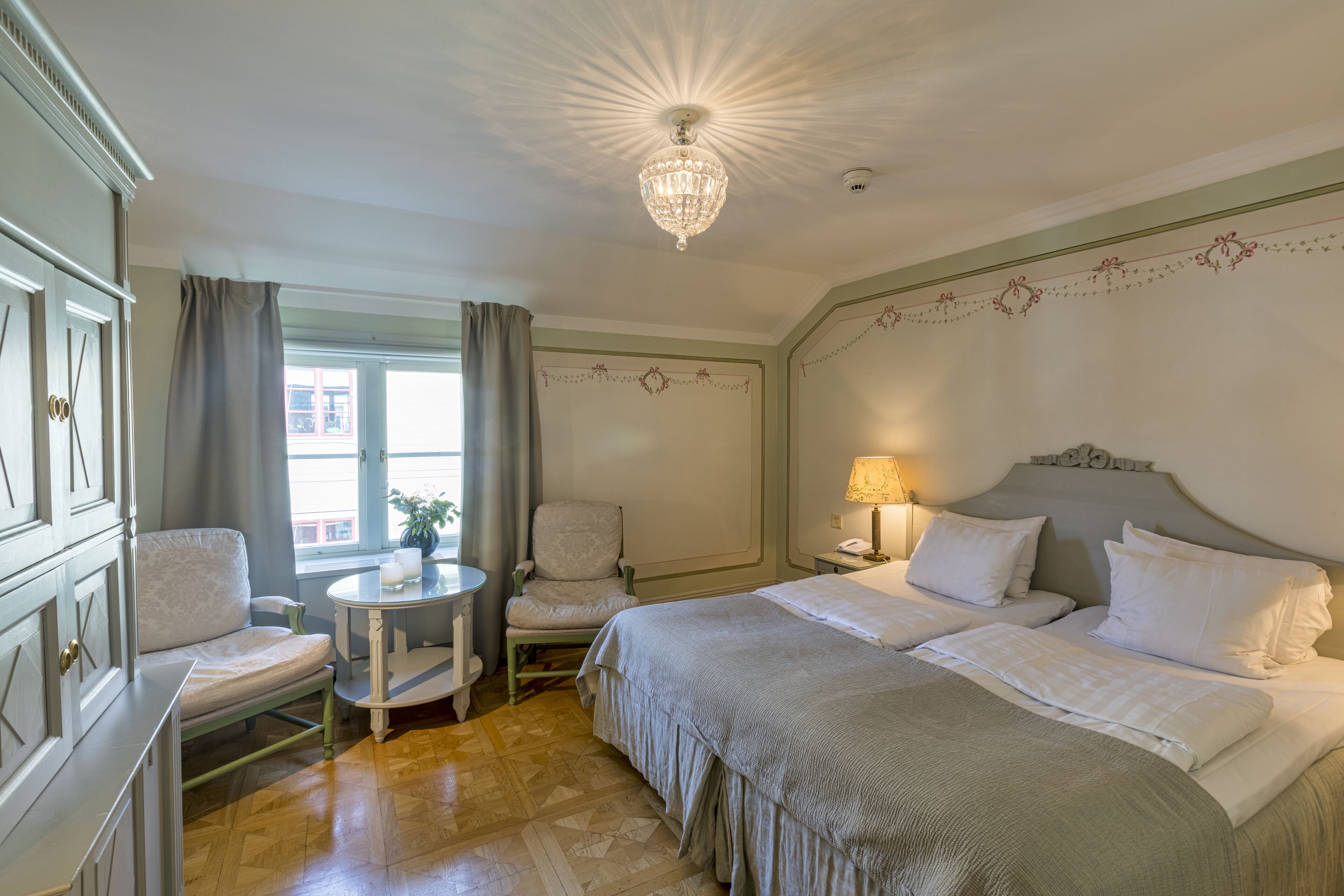 A hotel room with a large bed, two bedside tables, a chair, a small table, and a chandelier.