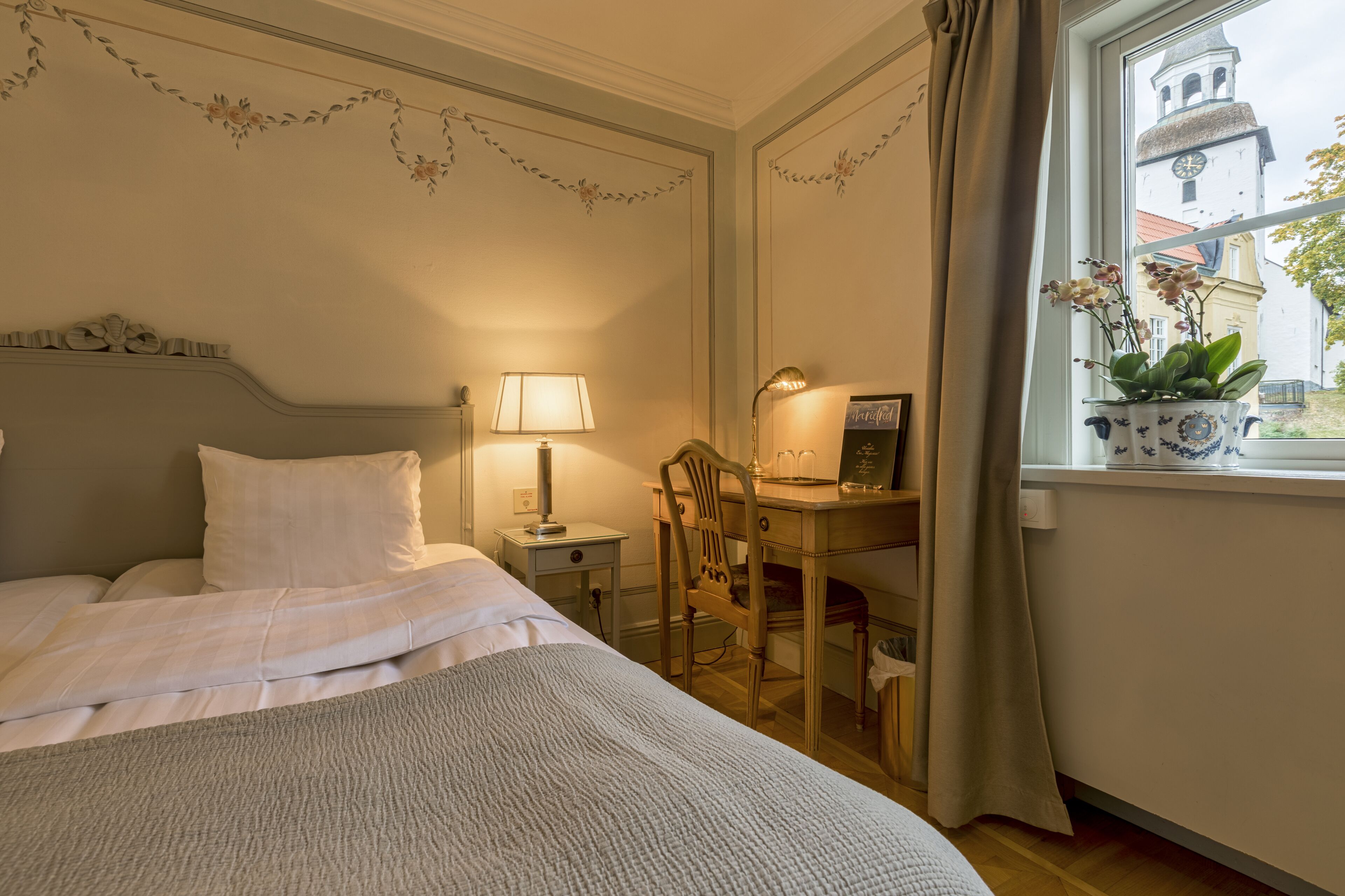 Ein ordentlich bezogenes Bett mit einer Lampe und einem Stuhl daneben. Ein Schreibtisch mit Lampe und einer Topfpflanze steht am Fensterbrett.