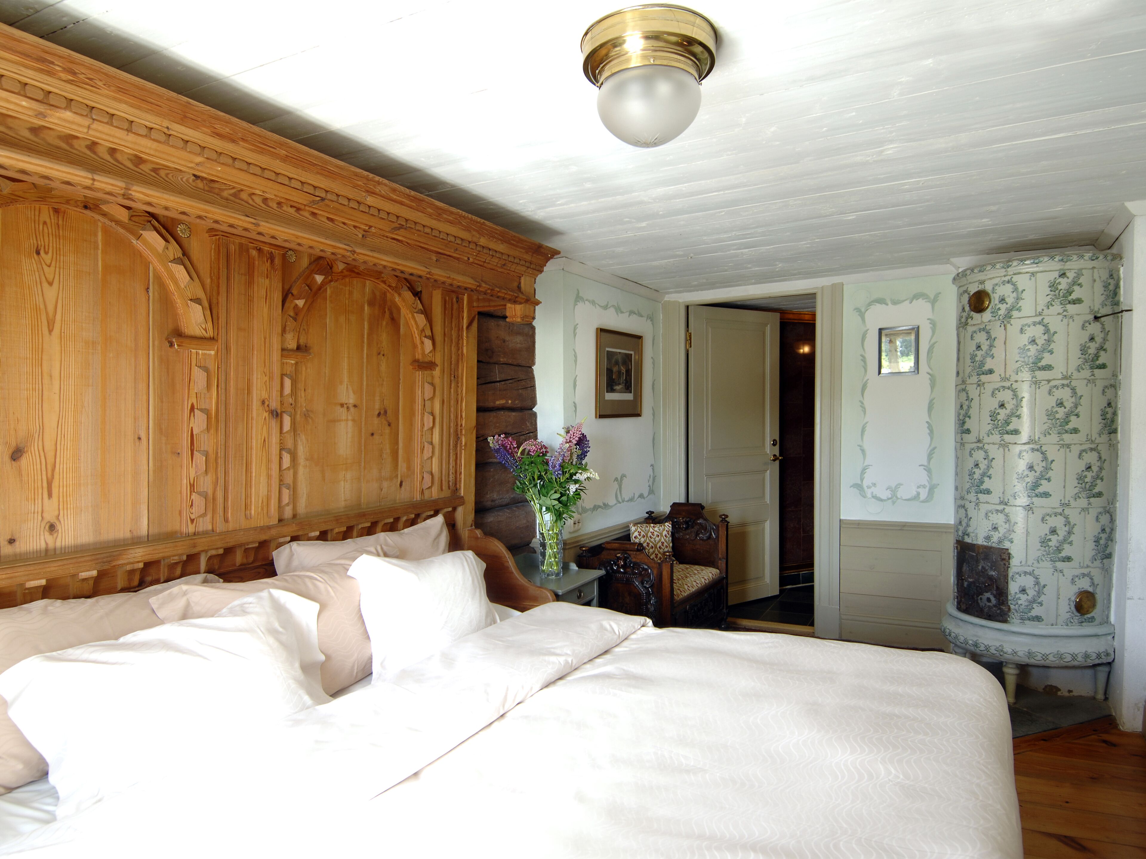 A bedroom with a large bed, wooden paneling on the walls, a vase with flowers, and a door leading to another room.