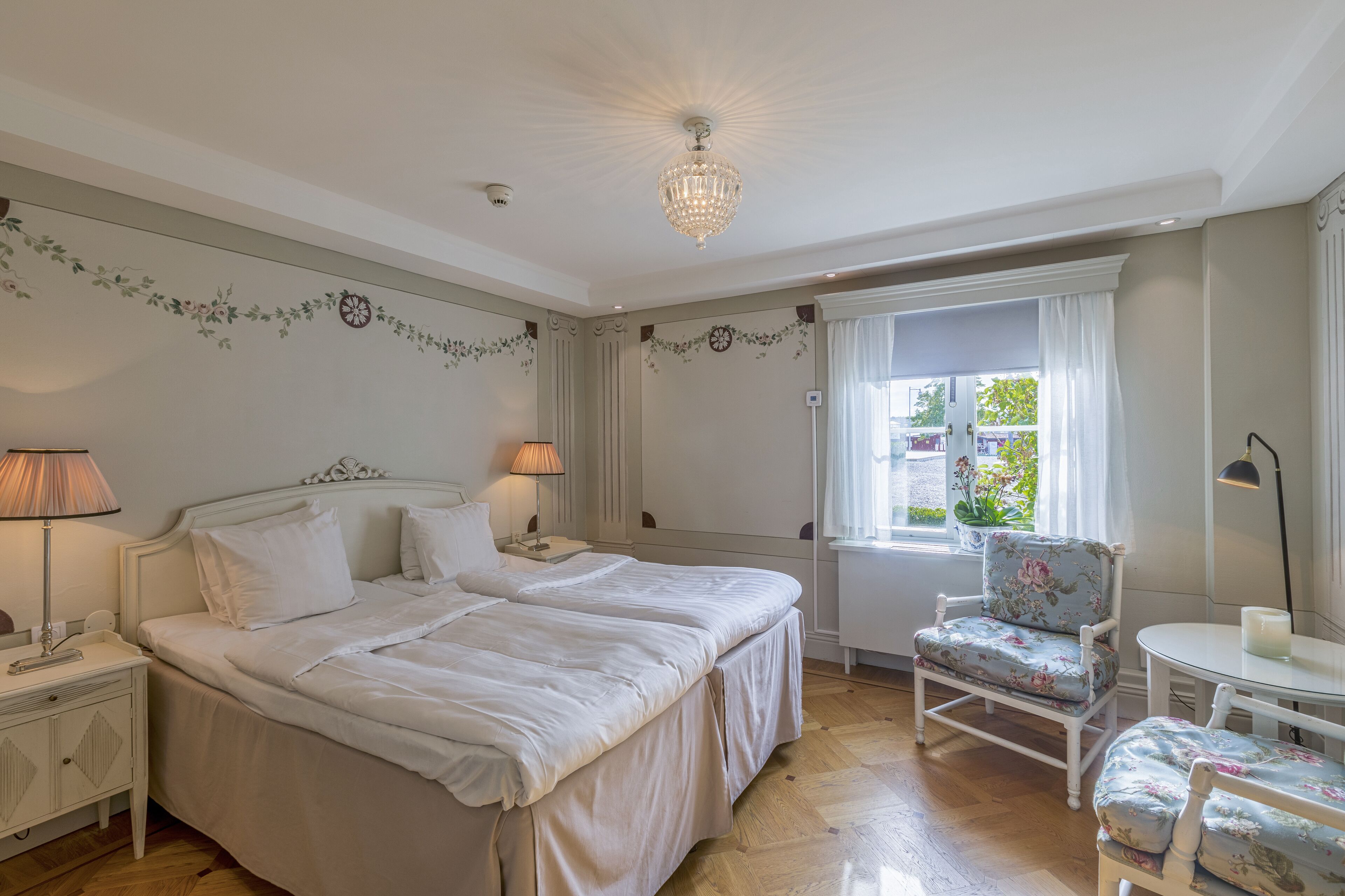 A bedroom with two beds, a nightstand, a chair, a table, and a chandelier.