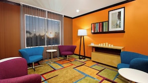 Habitación de hotel con un ventanal grande, una alfombra colorida, un sillón morado, un sillón azul, una mesa blanca y una lámpara de pie.