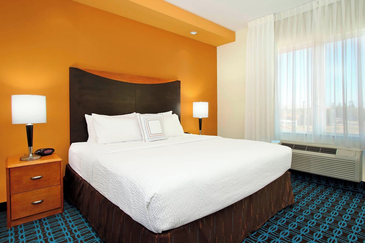 A hotel room with a large bed, two bedside tables with lamps, a window with sheer curtains, and a patterned carpet.