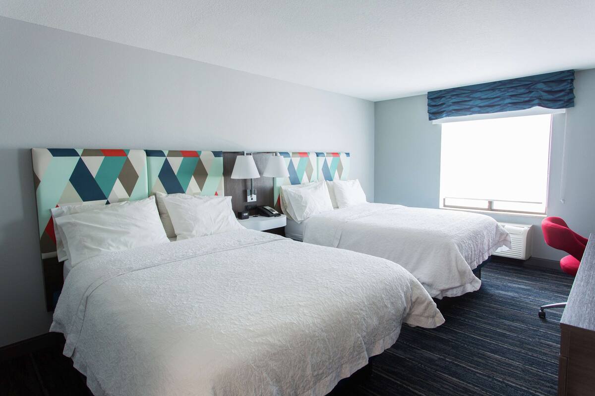 A hotel room with two beds, a desk, a chair, a window with a view, and a patterned headboard.