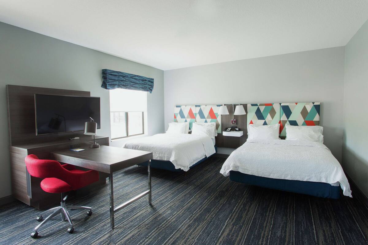 A hotel room with two beds, a desk with a chair, a TV, and a window with a blue curtain.