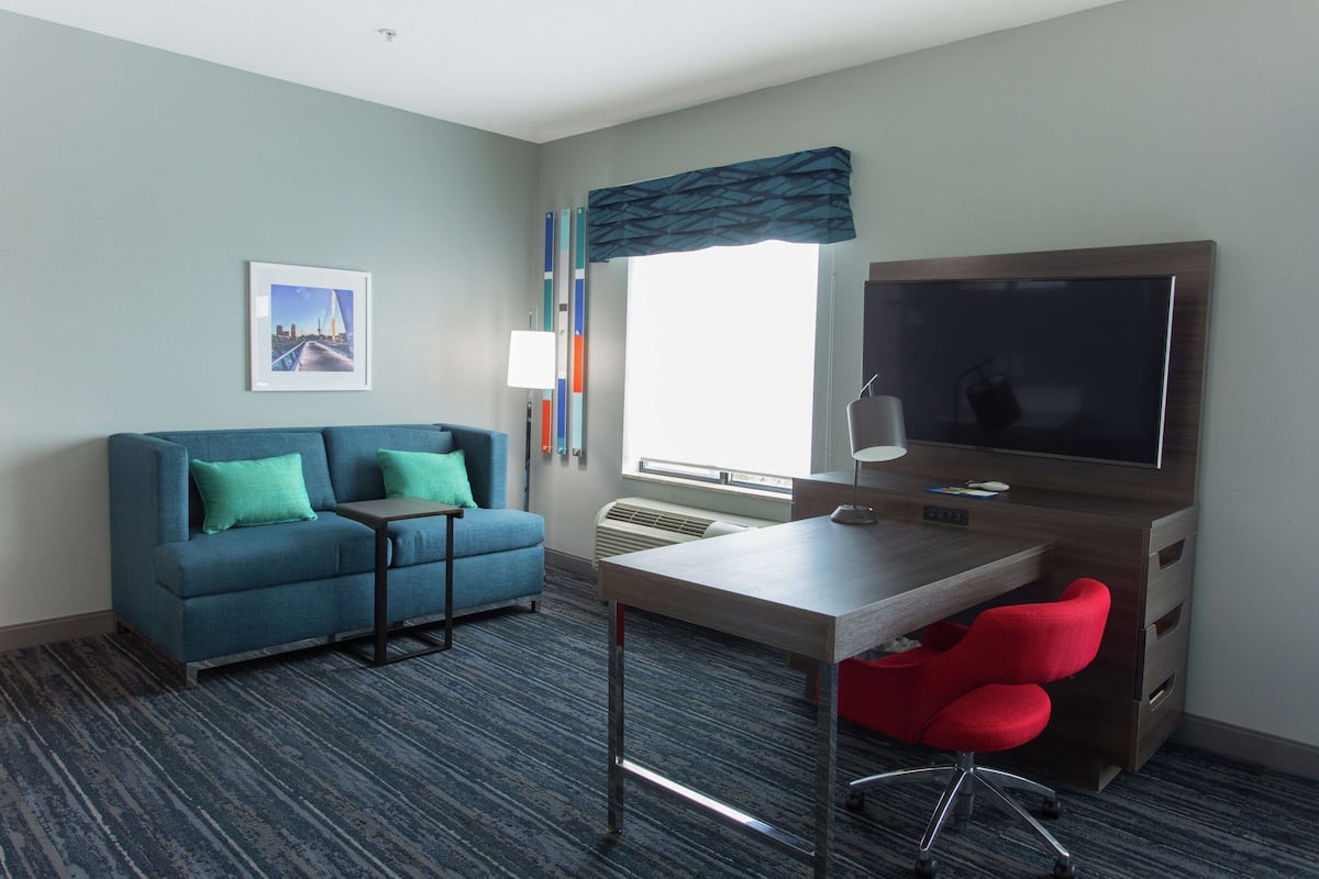 A hotel room with a blue sofa, a desk with a chair, a TV, and a window with a view of a bridge.