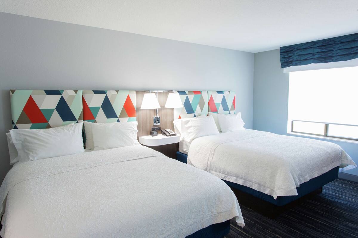 A hotel room with two beds, a nightstand with a lamp, and a geometric-patterned headboard.