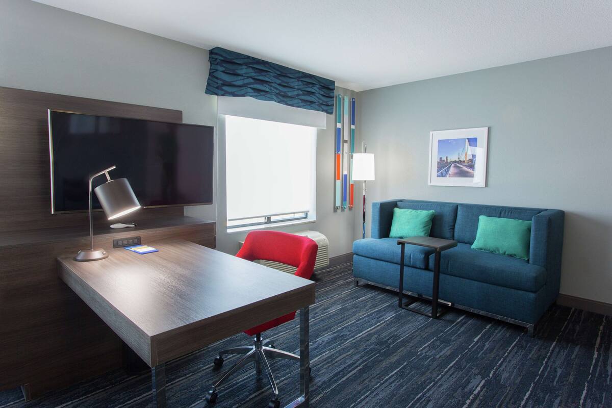 A hotel room with a desk, a red chair, a blue sofa, a flat-screen TV, a window with curtains, and a framed picture on the wall.