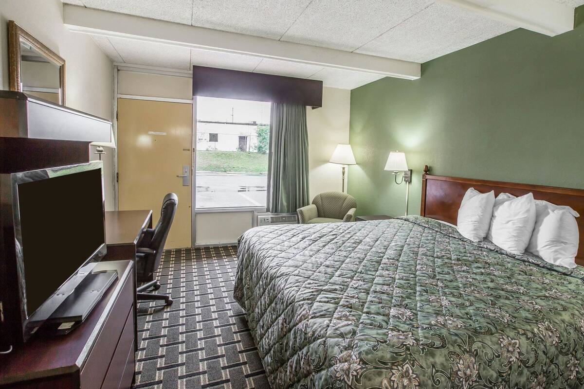 A hotel room with a bed, a desk with a television, a chair, a window, and a lamp.
