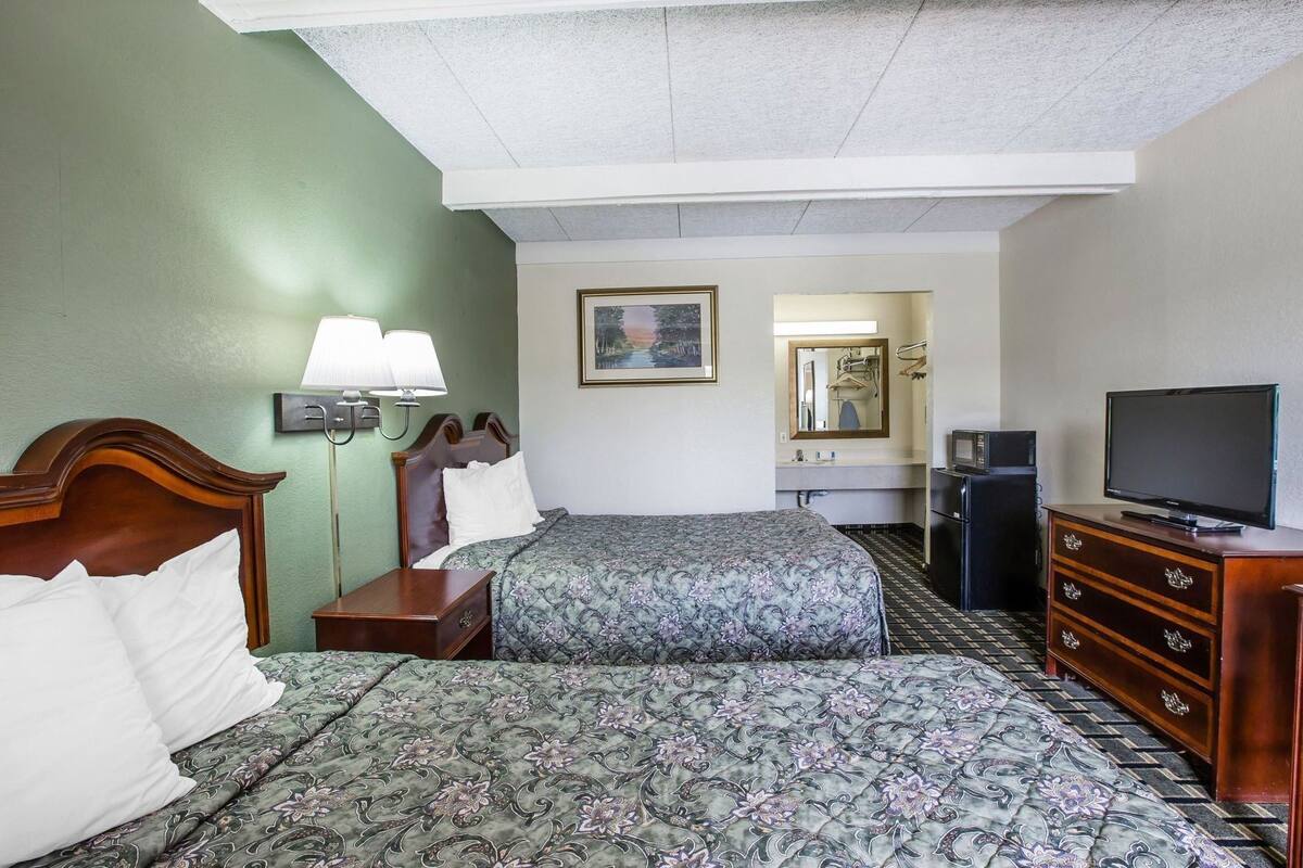 A hotel room with two beds, a TV, a dresser, a desk, a lamp, a mirror, and a painting on the wall.