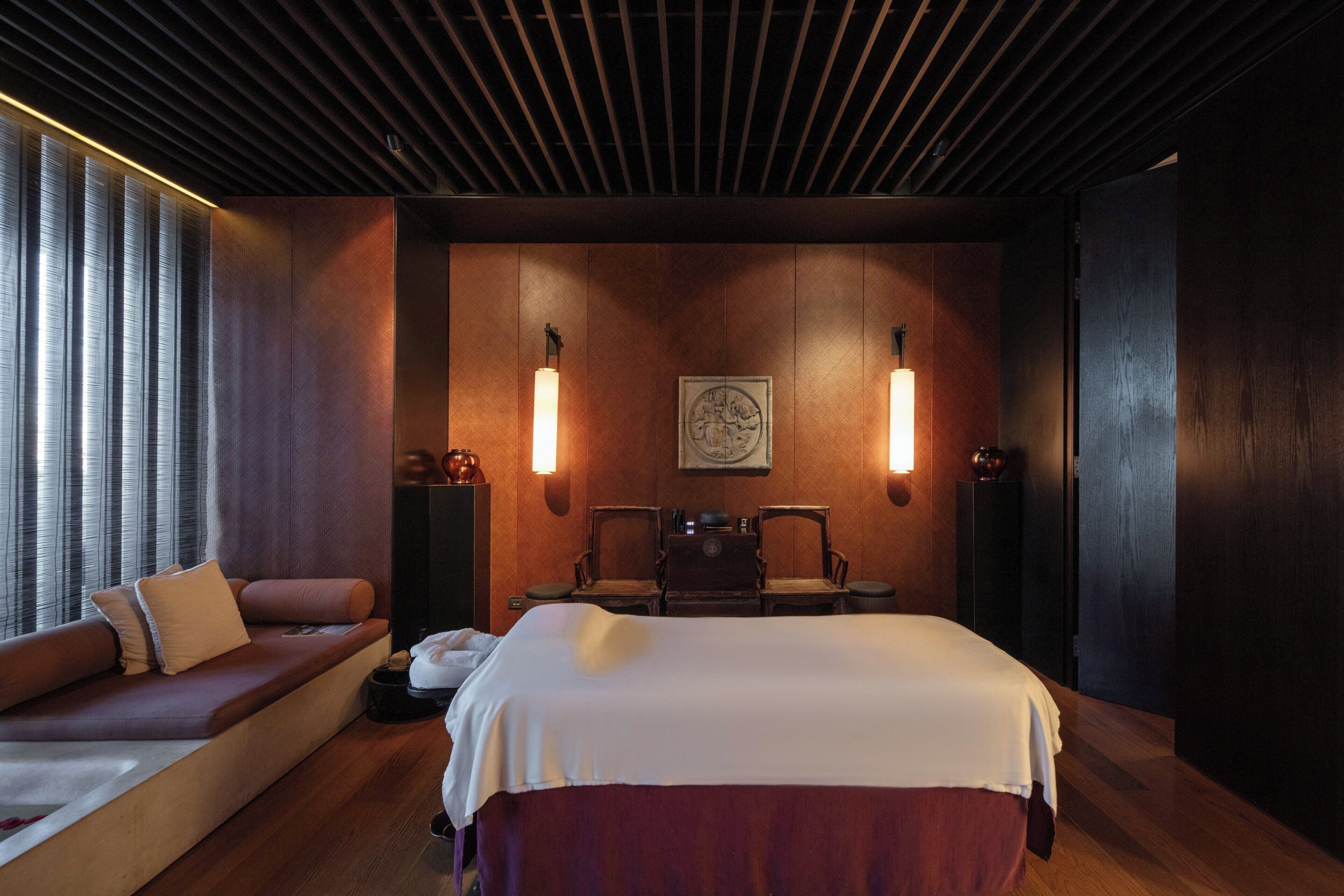 A modern hotel room with a large bed, a sofa, two chairs, a wooden ceiling, and wall-mounted lamps.