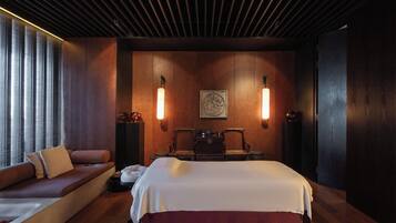 A modern hotel room with a large bed, a sofa, two chairs, a wooden ceiling, and wall-mounted lamps.