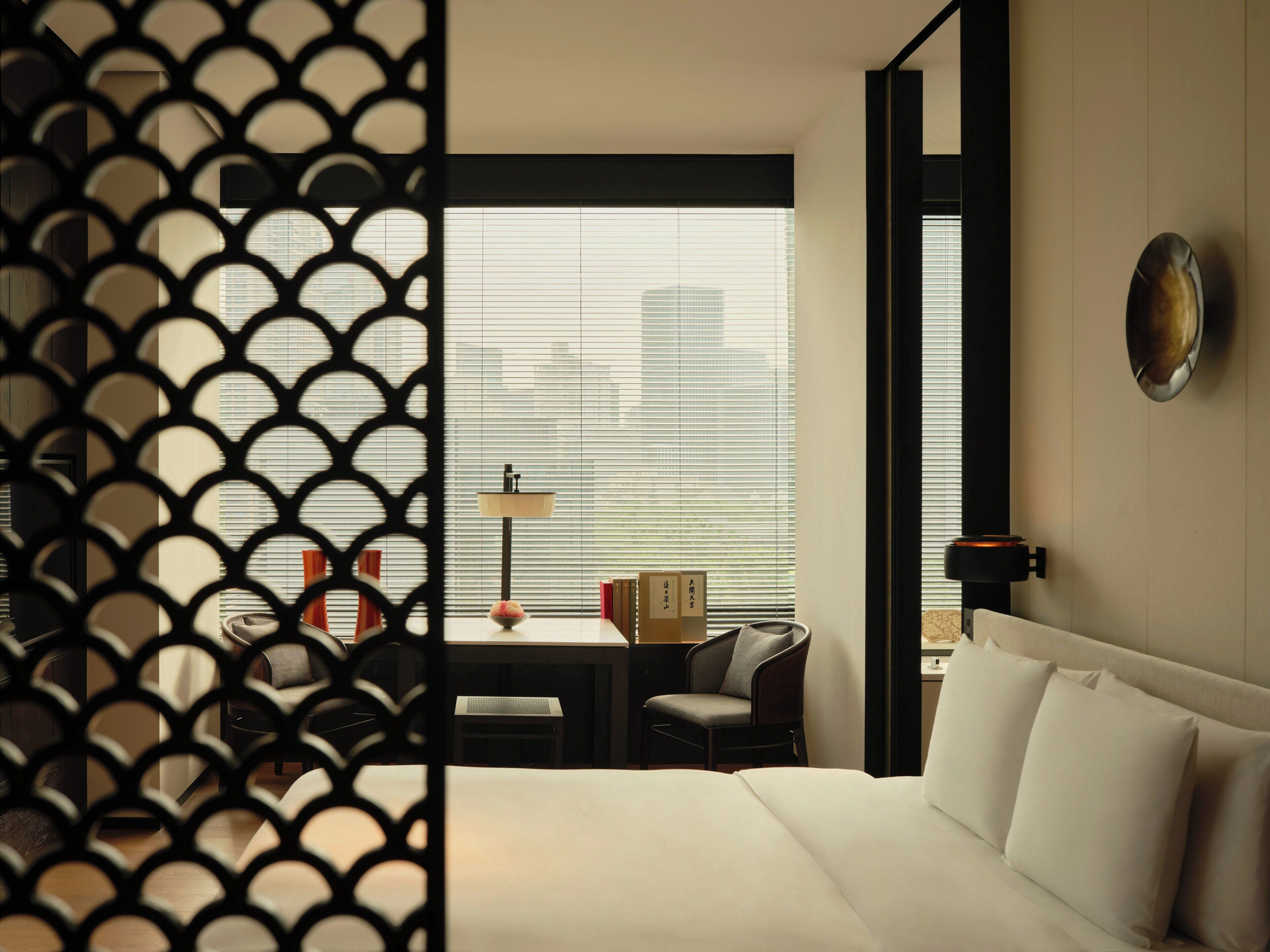 A hotel room with a bed, a desk, a chair, a lamp, and a view through the window.