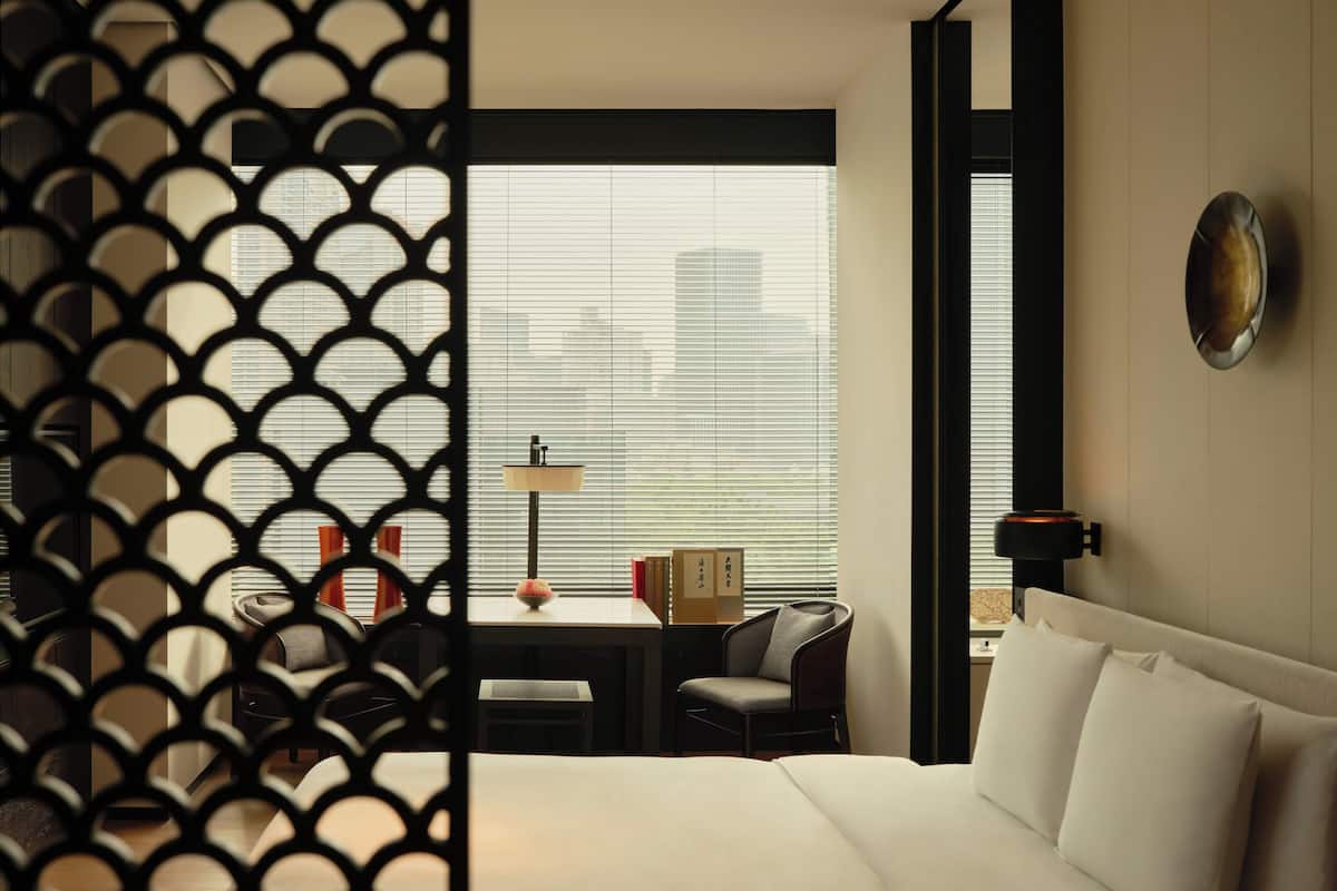 A hotel room with a bed, a desk, a chair, a lamp, and a view through the window.