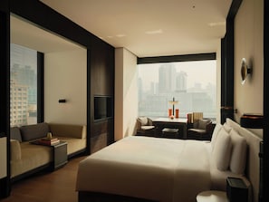 Una habitación de hotel moderna con una cama grande, un sofá, una mesa de comedor y vistas de la ciudad a través de amplios ventanales.
