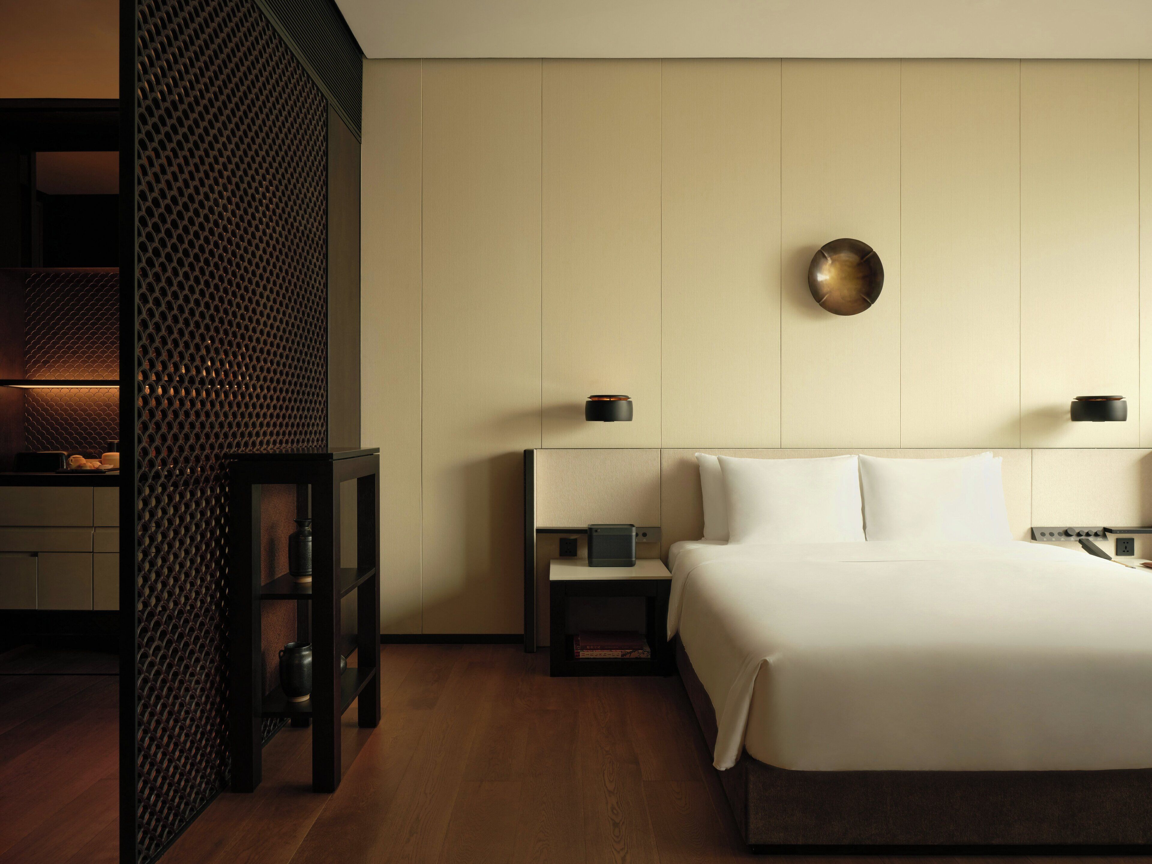 A hotel room with a bed, bedside tables, a wall-mounted lamp, and a textured partition.