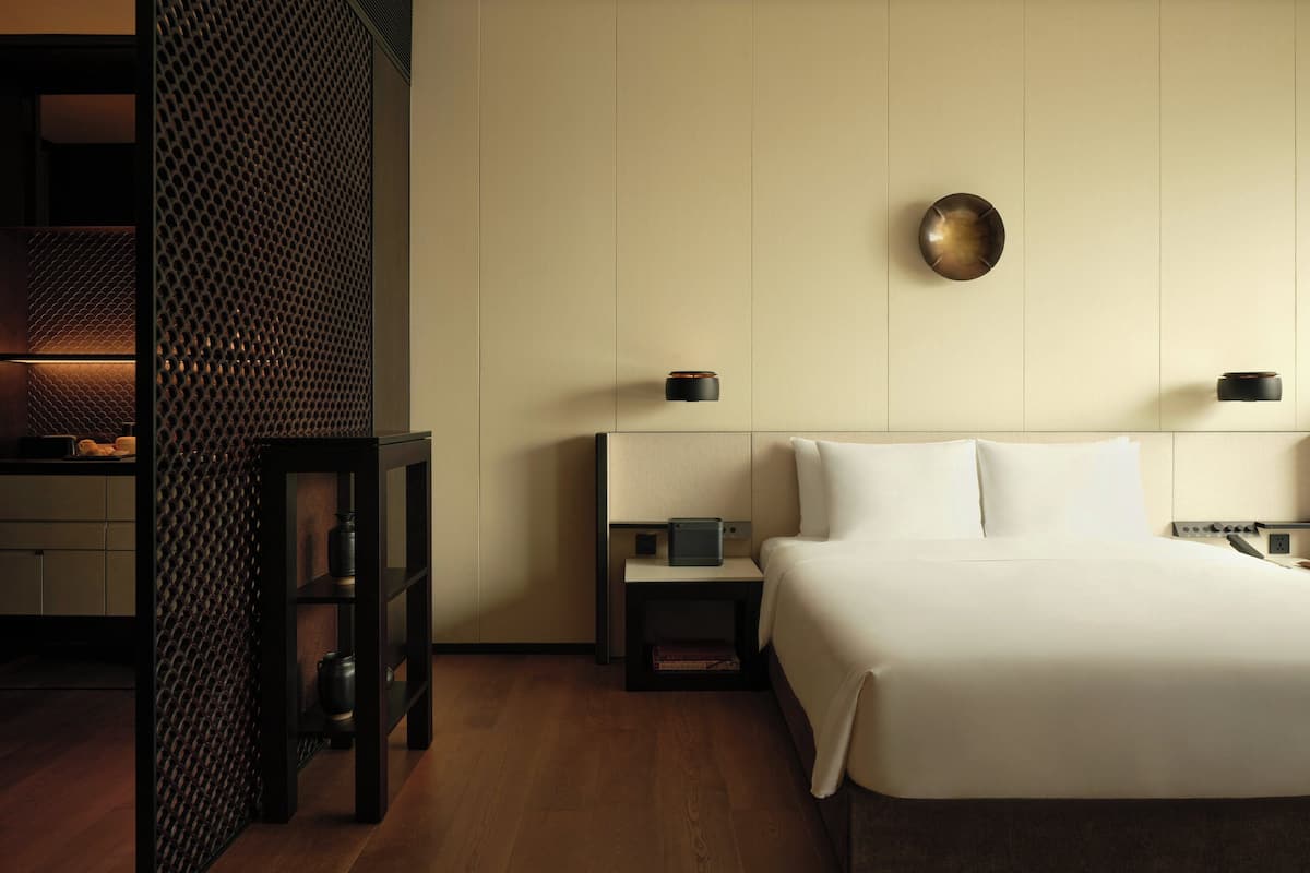 A hotel room with a bed, bedside tables, a wall-mounted lamp, and a textured partition.