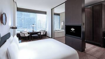 A modern hotel room with a large bed, a desk with a chair, a TV, and a view of the city through large windows.