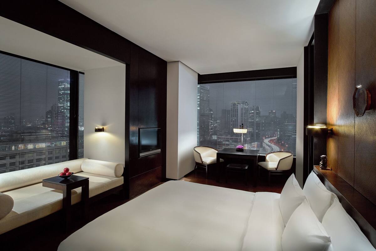 A modern hotel room with a large bed, a sofa, a small table, two chairs, and a view through large windows.