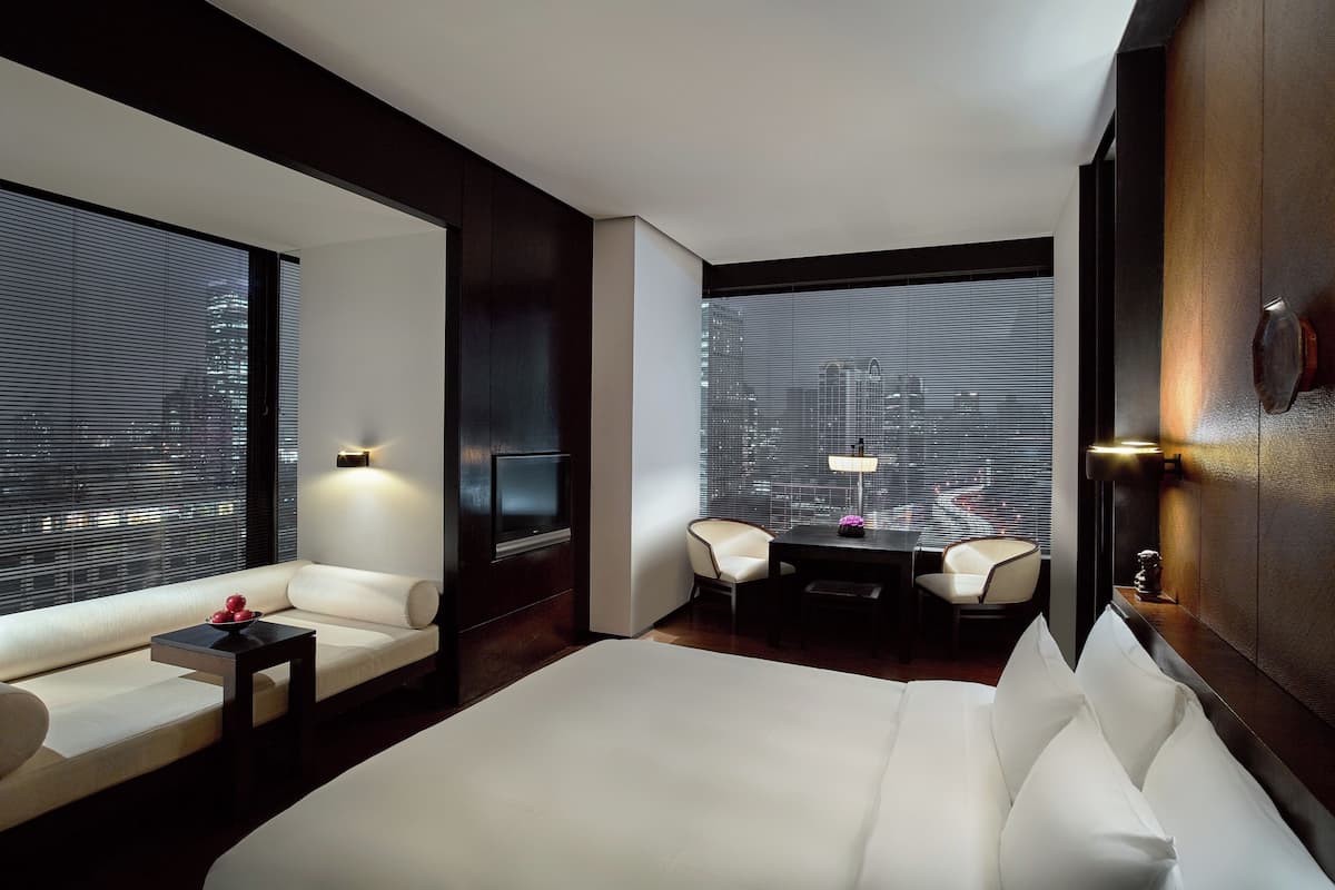 A modern hotel room with a large bed, a sofa, a small table, two chairs, and a view through large windows.