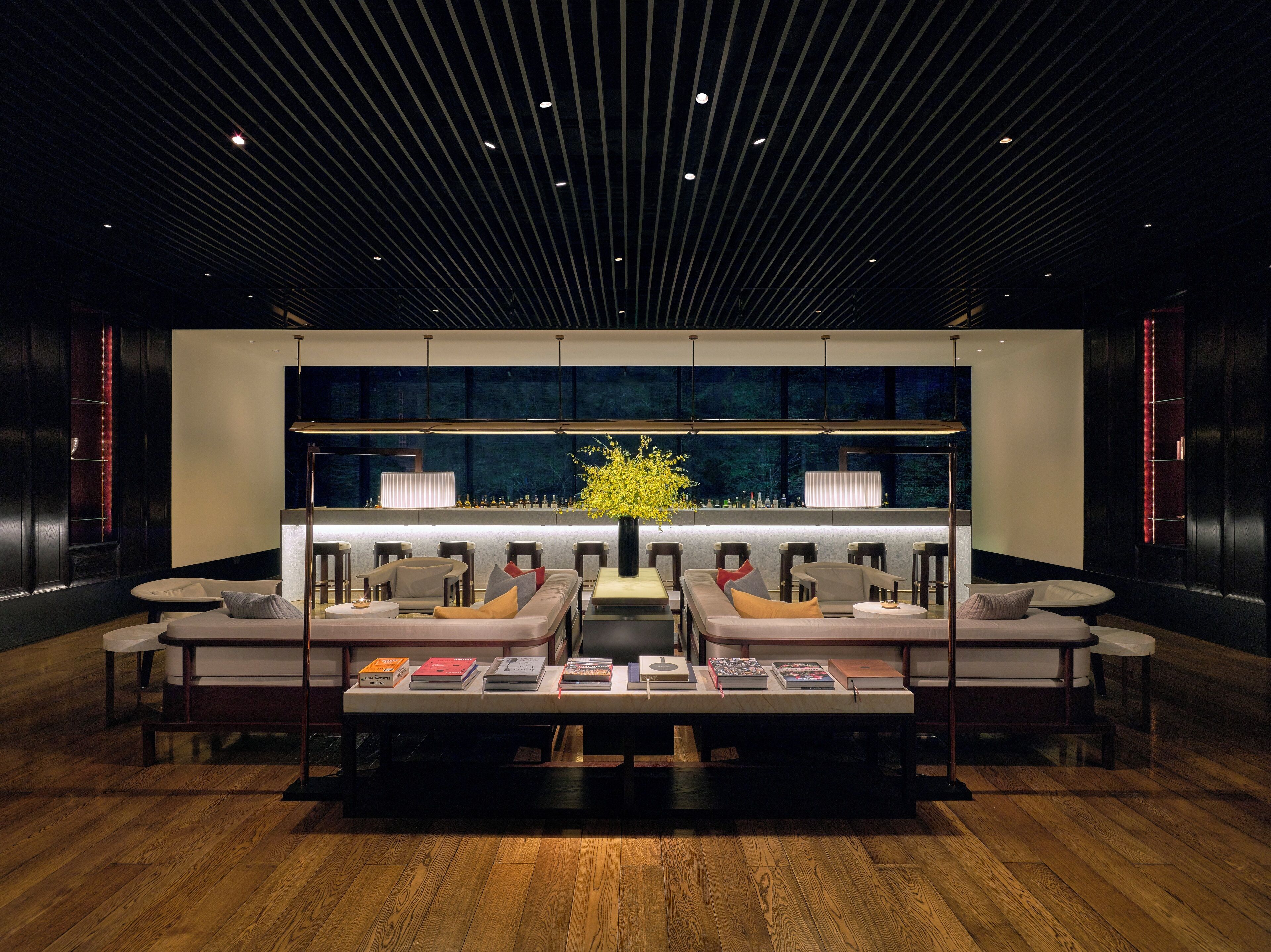A modern lounge area with a central seating arrangement, a bar with a large window, and a wooden floor.